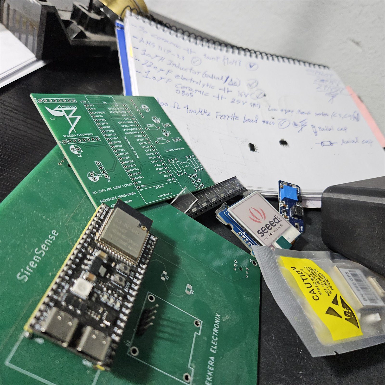 The green SirenSense transponder circuit board beside a long-range radio module, an ESP32 board and a handwritten list of power components