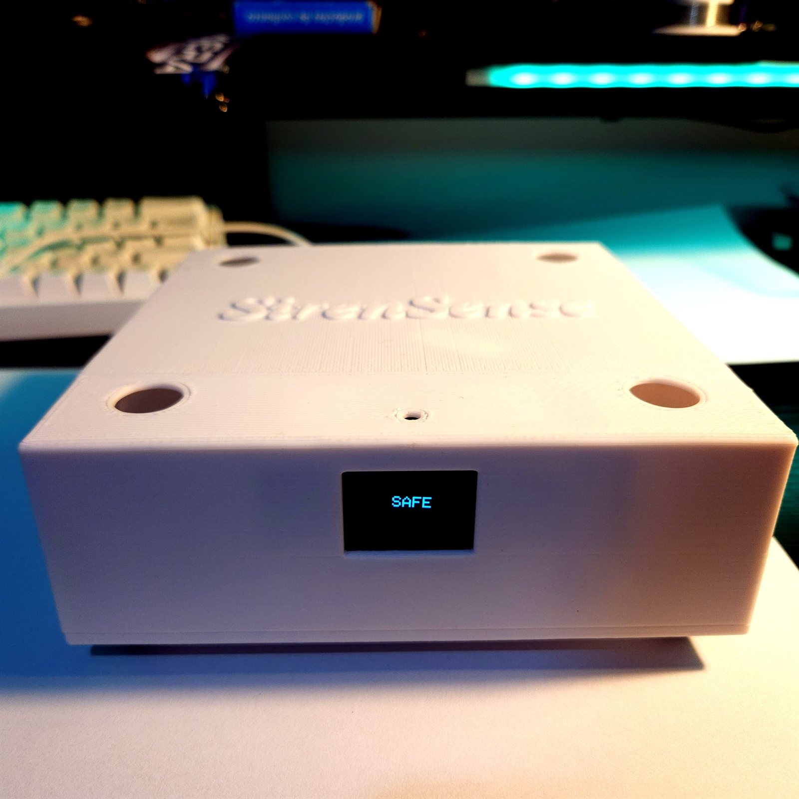 A white printed enclosure with SirenSense embossed on the lid, four circular microphone ports and an OLED showing SAFE