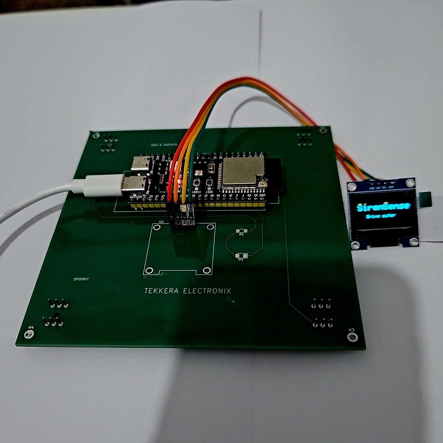 The REV 1 board on a bench with an OLED breakout displaying the SirenSense boot screen reading Oye antes