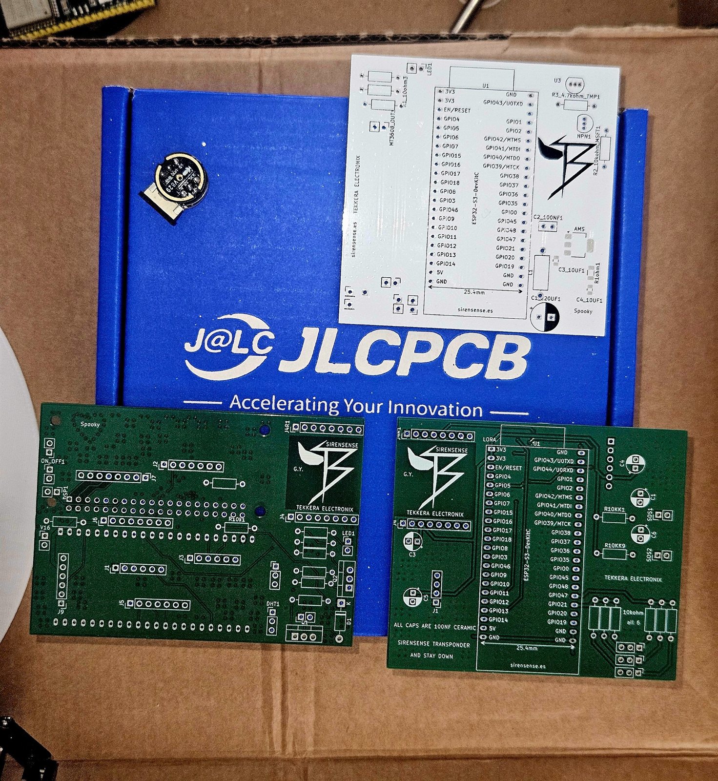 Three green fabricated circuit boards on a JLCPCB box, showing TEKKERA ELECTRONIX and SirenSense silkscreen