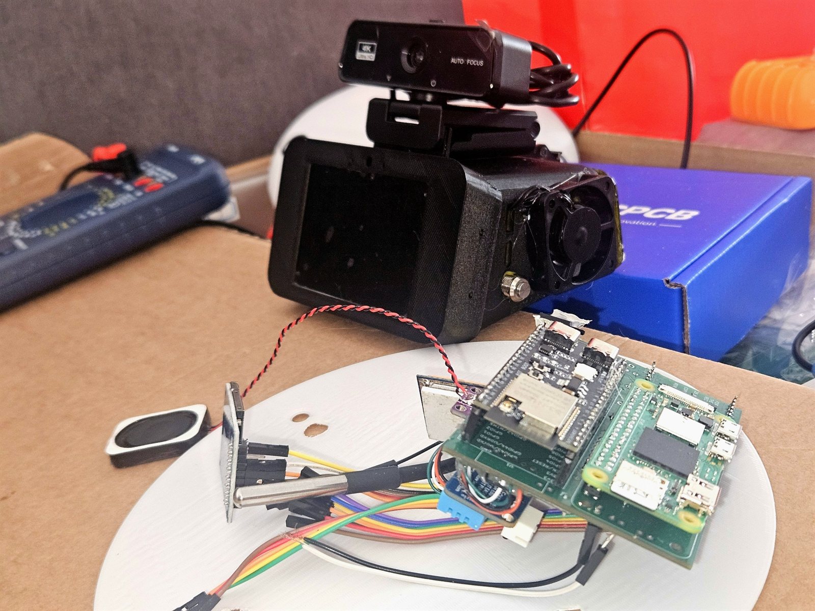 Bench test setup with a camera module, OLED, stacked green boards, a multimeter and a printed disc